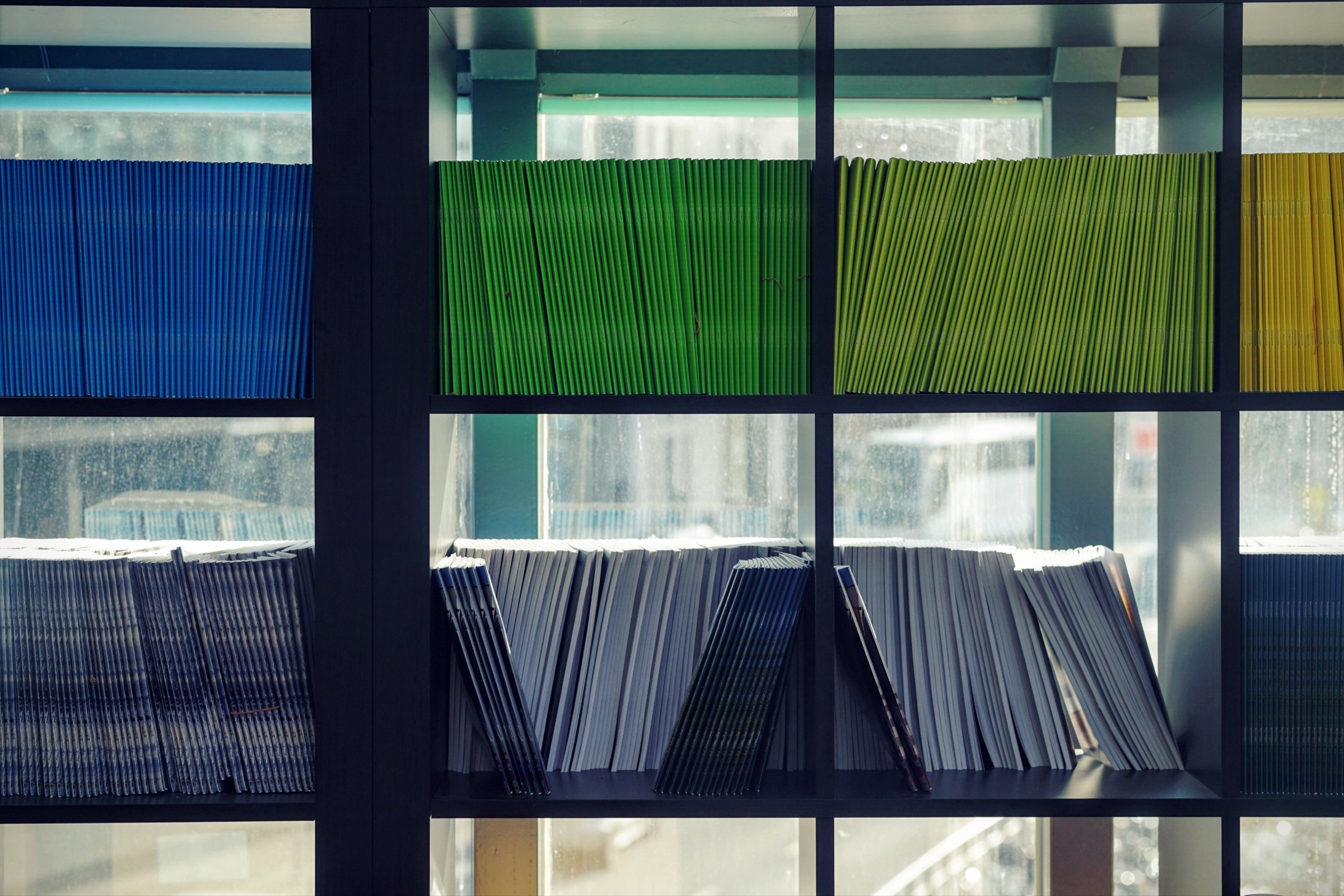 Een kast vol gestapelde magazines en agenda’s in blauw, groen en geel, met daglicht erachter.