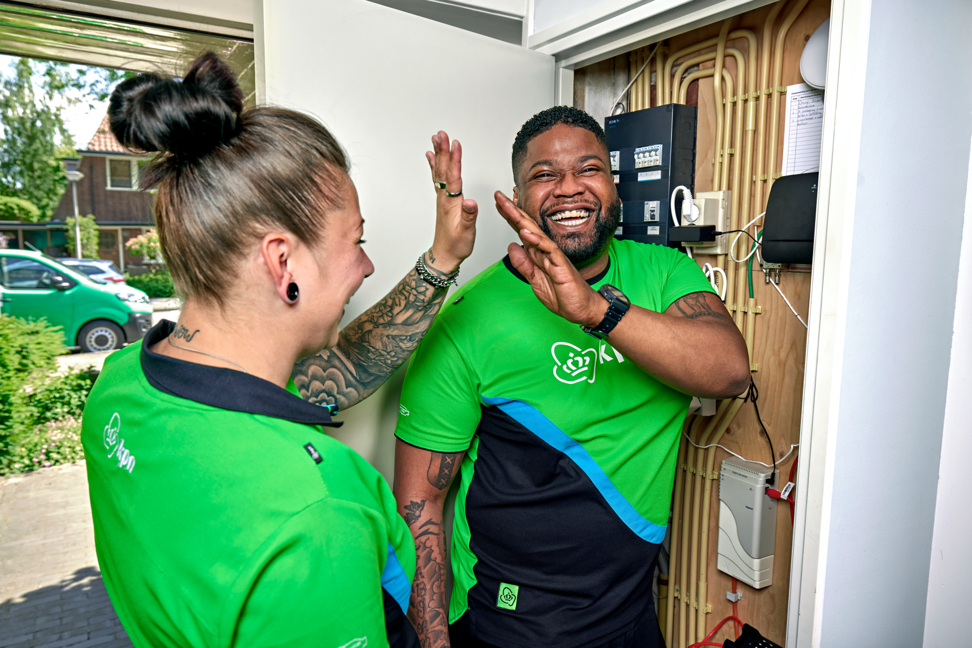 Twee KPN-monteurs in groene bedrijfskleding geven elkaar lachend een high five voor een geopende meterkast.
