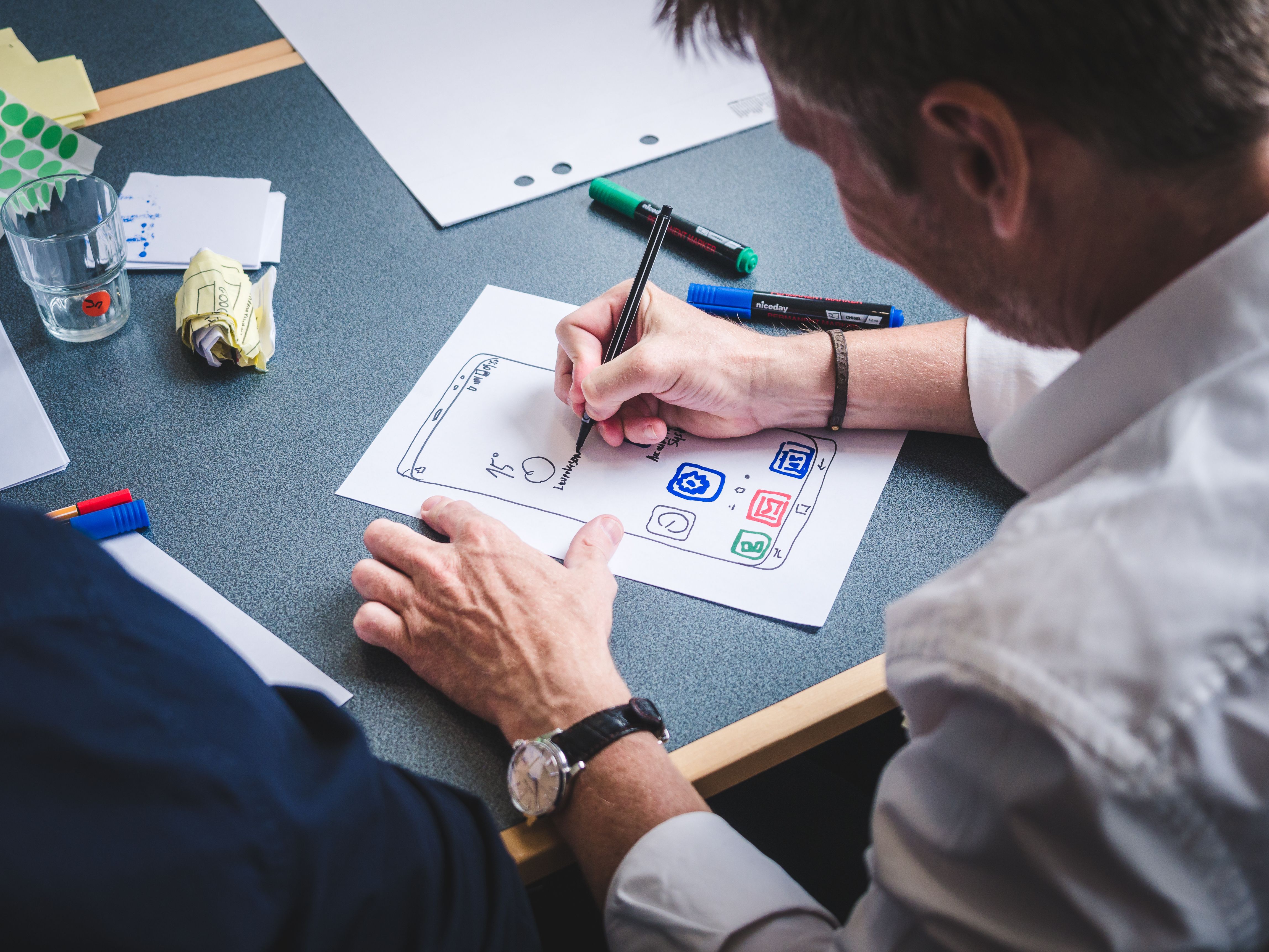 Een ontwerper schetst met stift een app-scherm op papier, tussen markers en post-its.